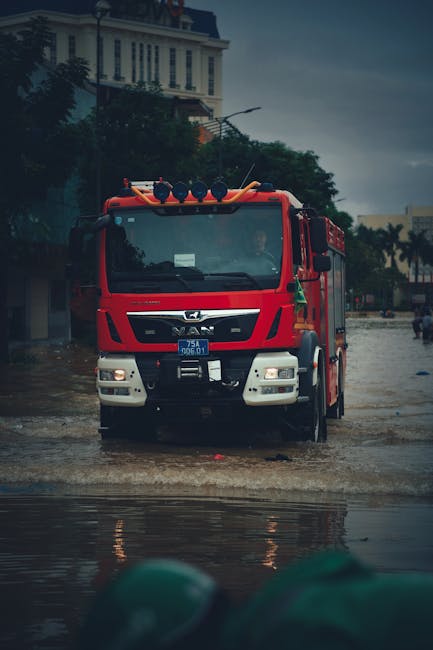 A group of emergency responders and cleaning personnel in yellow and orange raincoats working on a wet, flood-affected residential street in Clapham Junction. The scene includes a white van parked in 