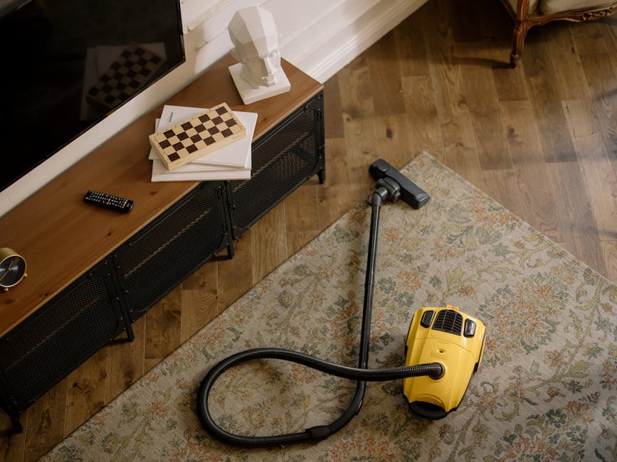 An elderly man with grey hair and a grey long-sleeve shirt is using a vacuum cleaner to perform surface cleaning in a bright, modern living room. The room features a beige sofa with blue and orange cu